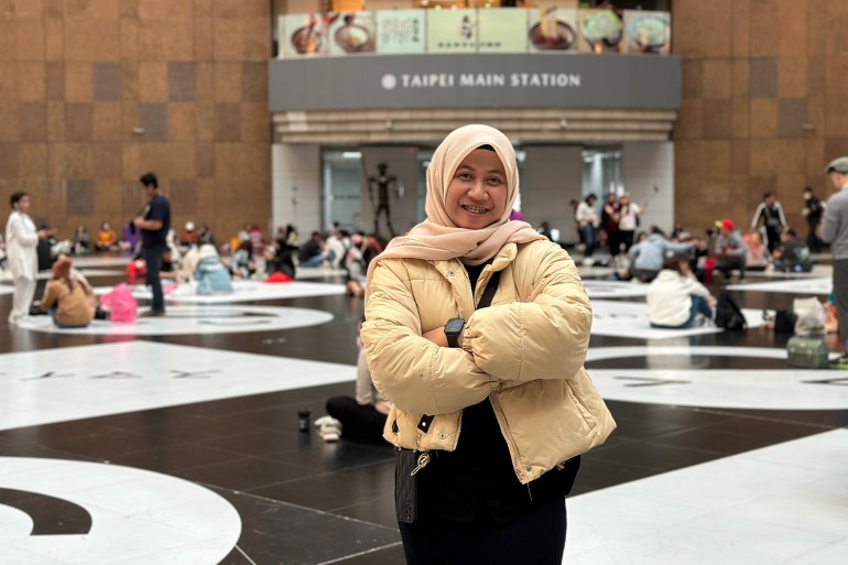 Anggi Sofiasyah Lacuba. Está en la estación de Taipei. Lleva una chaqueta con un pañuelo en la cabeza y tiene los brazos cruzados.